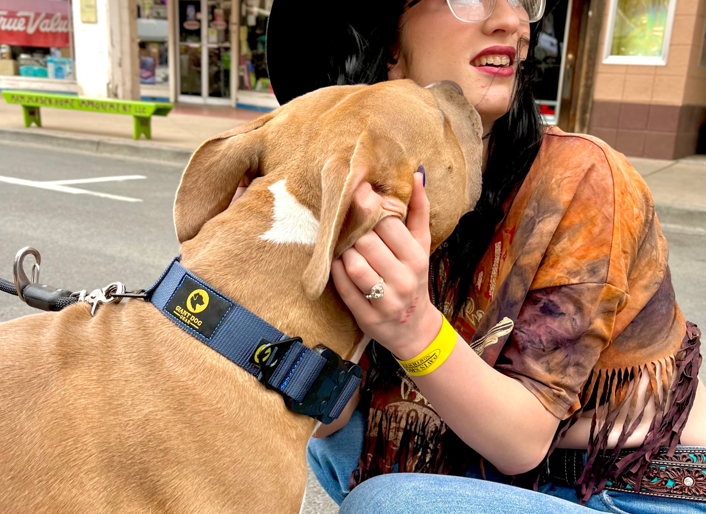 Woman in a black cowboy hat at a street fair petting a Pit Bull mix dog wearing a Giant Dog Gear navy blue collar and No-Pull Wrap Leash with American Flag Fourth of July decorations on a store in the background